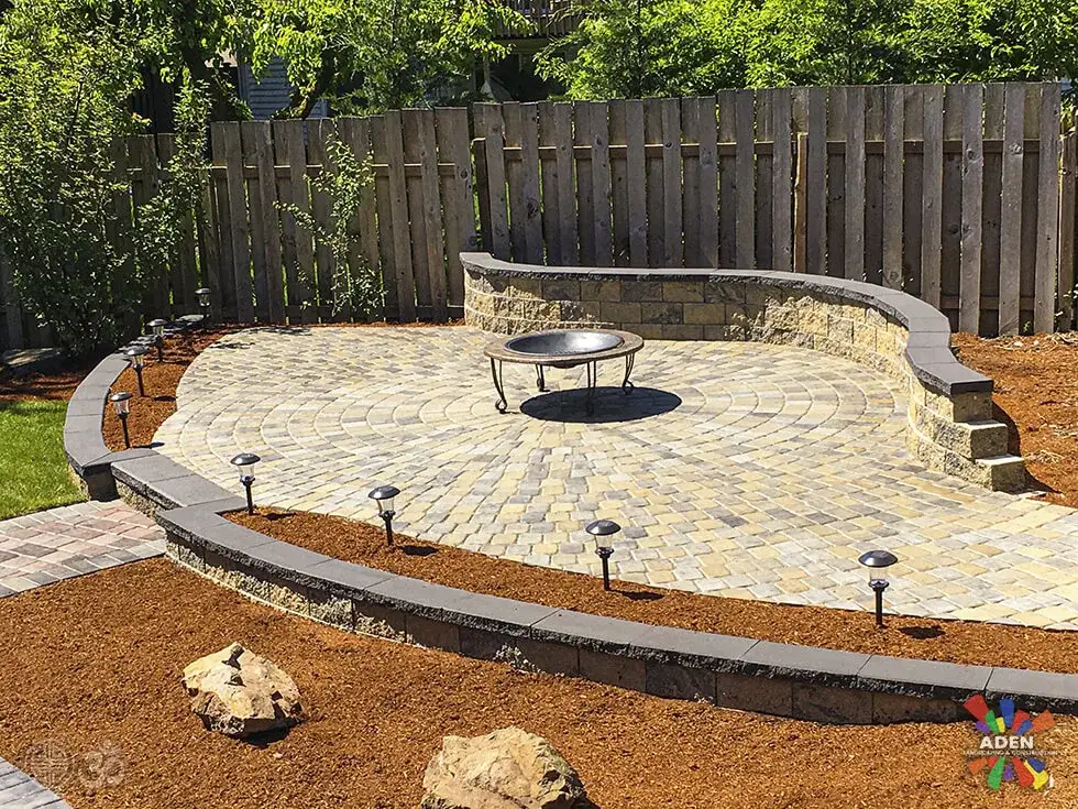 Circular paver terrace with a curved seat wall, a central fire bowl and path lights, surrounded by fresh bark mulch