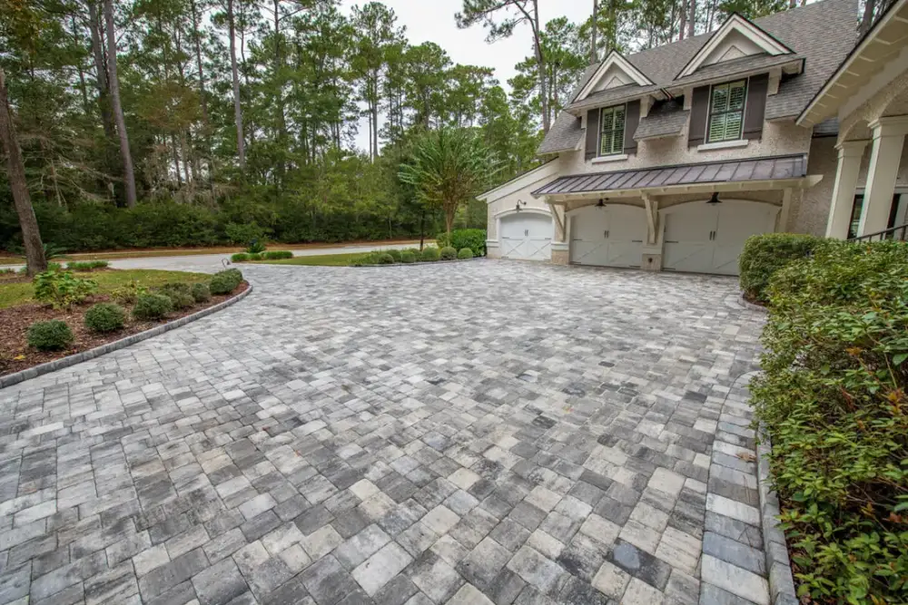 Wide grey paver driveway curving up to the three-car garage of a large house surrounded by conifers