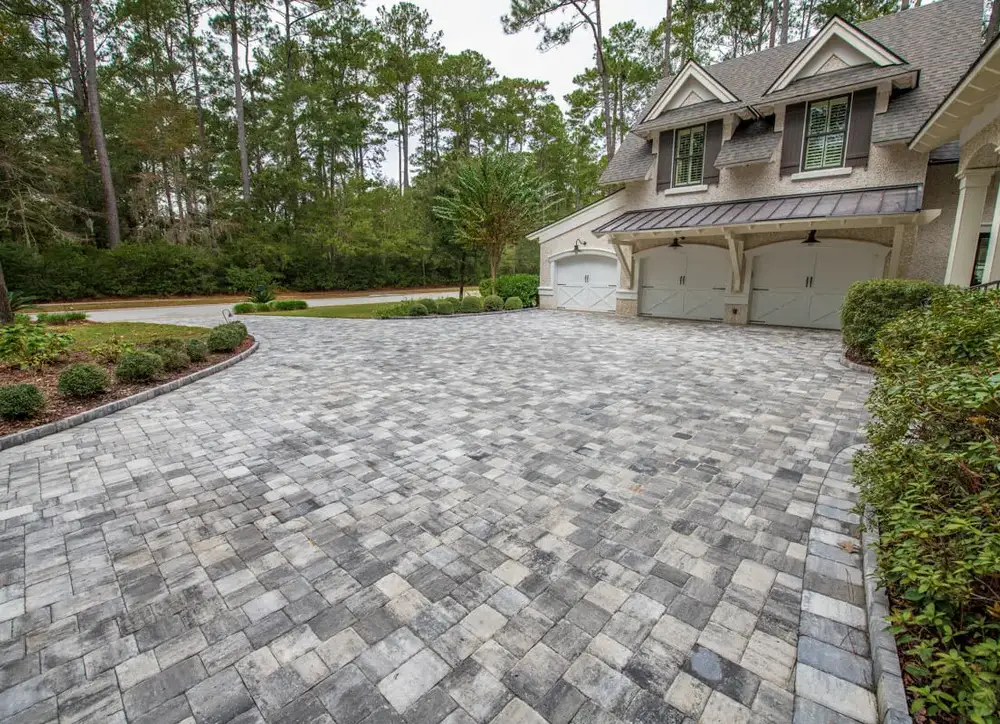Wide grey paver driveway curving up to the three-car garage of a large house surrounded by conifers