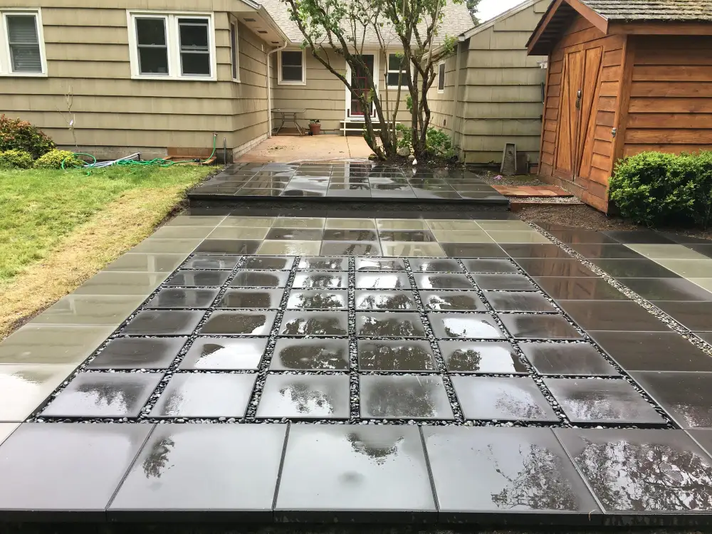 Grid of large charcoal pavers with grass growing in the open joints, wet from rain, beside a house and a cedar shed
