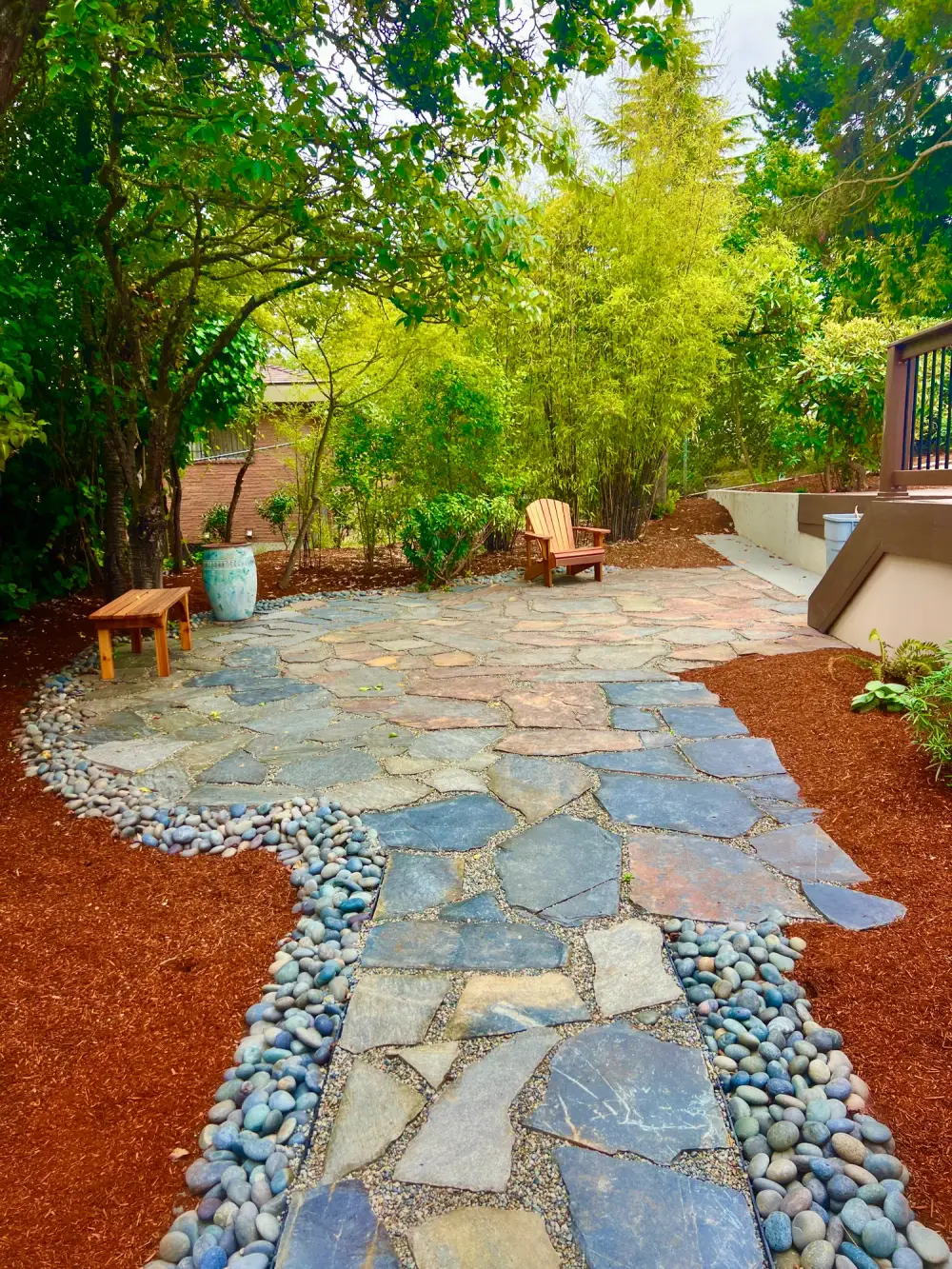 Irregular flagstone terrace with tight joints, bordered by washed river rock and red bark mulch under mature trees