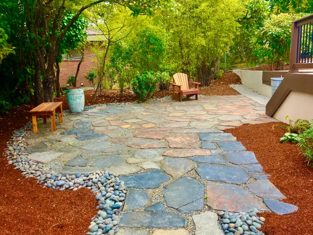 Irregular flagstone terrace with tight joints, bordered by washed river rock and red bark mulch under mature trees