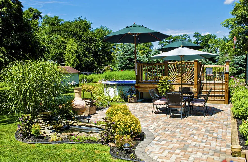 Tumbled paver patio with a dining set under green umbrellas beside a planted pond and a raised timber gazebo deck