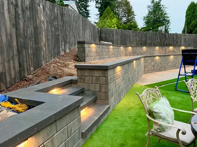 Block retaining wall and seat wall at dusk with recessed lighting glowing in the wall face and step risers, beside an artificial turf lawn