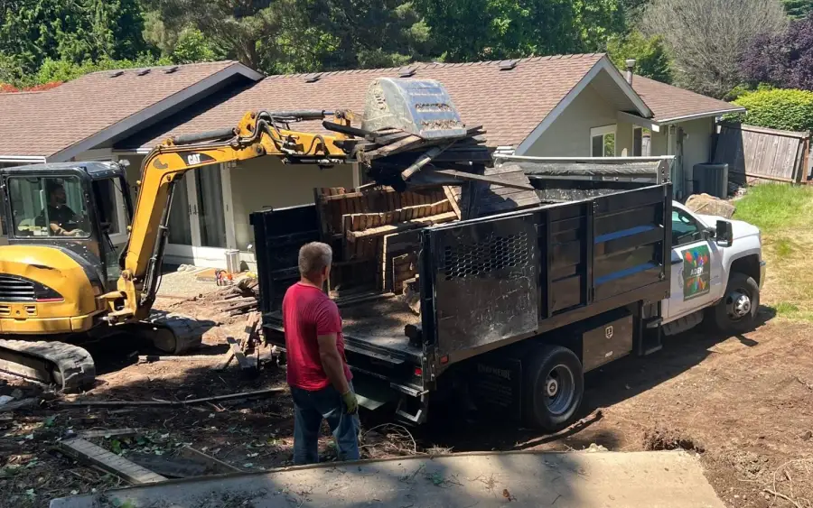 Excavator loading cleared timber and vegetation into an Aden Landscaping dump truck on a Seattle property