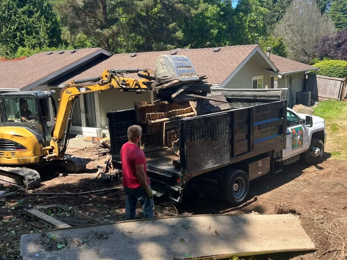 Excavator loading cleared timber and vegetation into an Aden Landscaping dump truck on a Seattle property