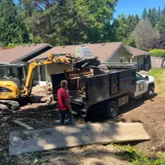 Excavator loading cleared timber and vegetation into an Aden Landscaping dump truck on a Seattle property