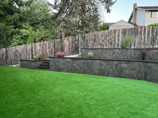 The same block retaining wall in daylight, running the length of a cedar fence above a finished artificial turf lawn