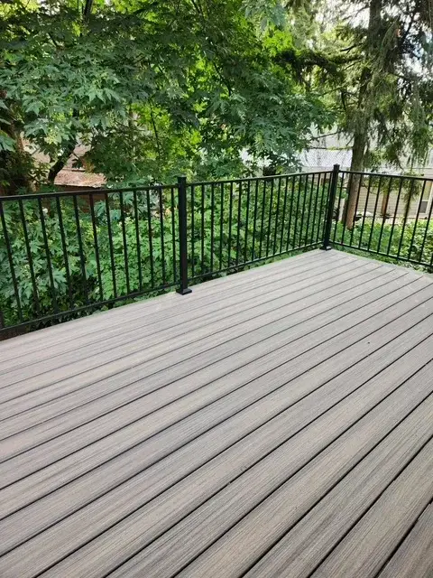 Composite deck in grey and brown tones with black metal railing, surrounded by mature broadleaf trees in a Seward Park garden