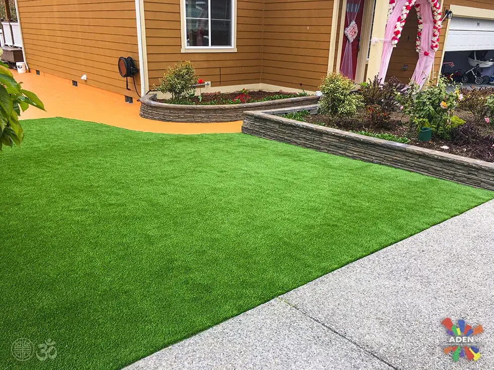 Artificial turf front lawn meeting a curved block retaining wall and a concrete path beside a tan house