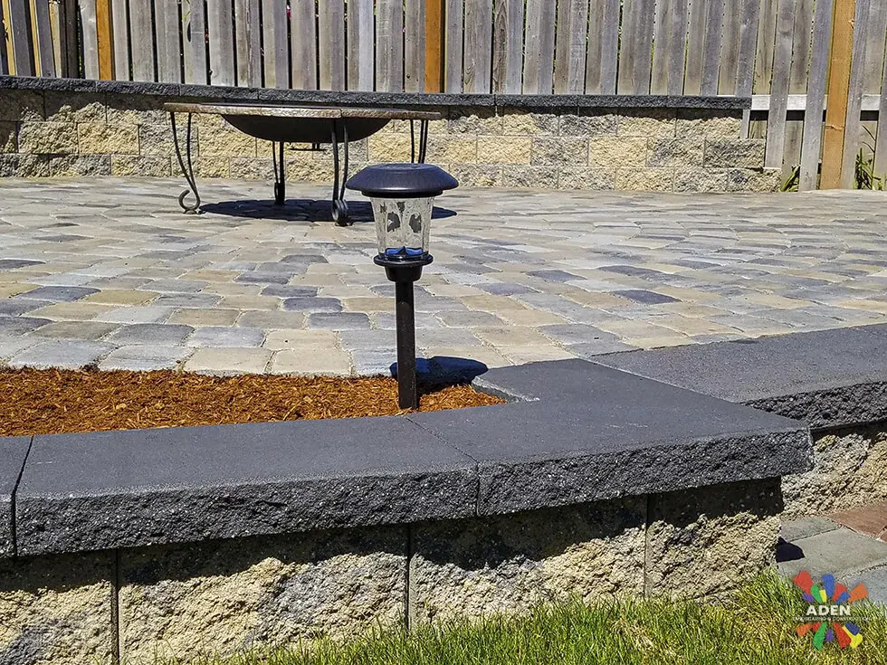 The same raised paver terrace seen from the lawn, with a black fire bowl on the paved surface and a path light in the foreground