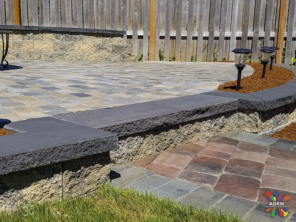Raised paver patio with a dark charcoal cap course, path lights along the edge and a bark bed below it
