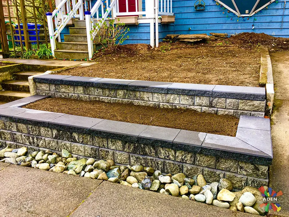 Two tiers of grey block retaining wall with concrete steps between them and a band of river cobble along the base