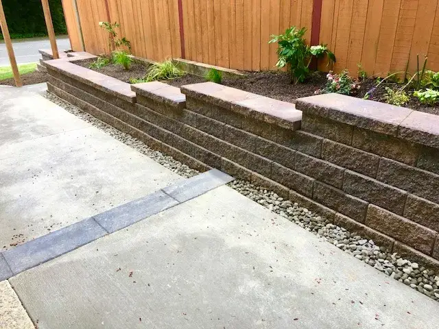 Block retaining wall with a brick-red capping course and a clean rock drainage strip between the wall and a poured concrete patio