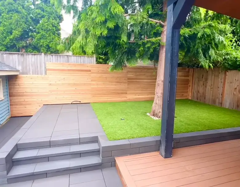 Seattle back garden with an artificial turf lawn laid around a mature conifer, a grey slab terrace with wide steps, horizontal cedar fencing and a covered deck on dark posts