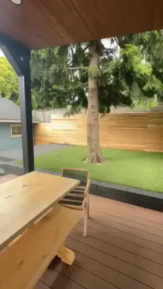 View from under a covered deck in Seattle, past a timber table and chair, out to an artificial turf lawn and the conifer growing through it