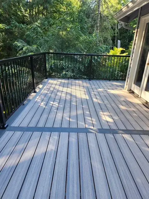Grey composite deck with a darker inlaid border board and black metal railing, backing onto conifers at a Wedgwood house