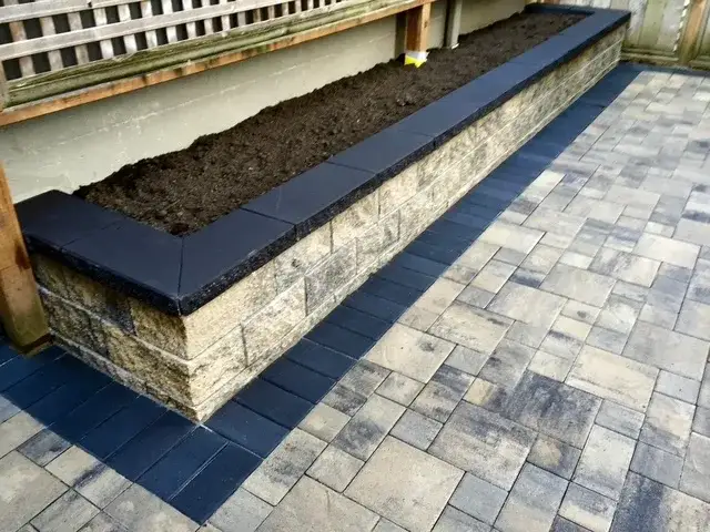 Long raised planter of split-face block with a charcoal capping course, filled with fresh soil, on a two-tone paver terrace
