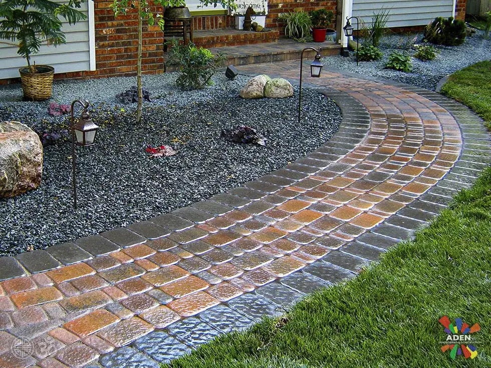 Curved brick paver walkway with a cobble edge and low landscape lights, crossing a bed of dark gravel towards a lawn