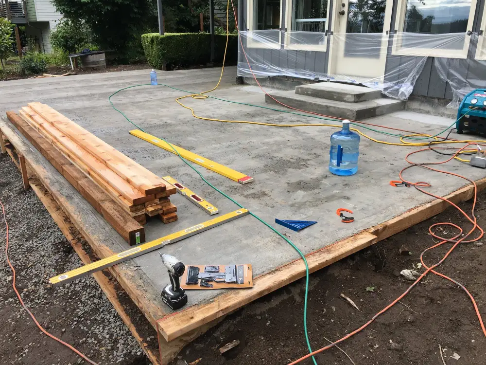 Timber joists and boards laid out on a concrete slab beside a house with hoses and tools, at the start of a deck build