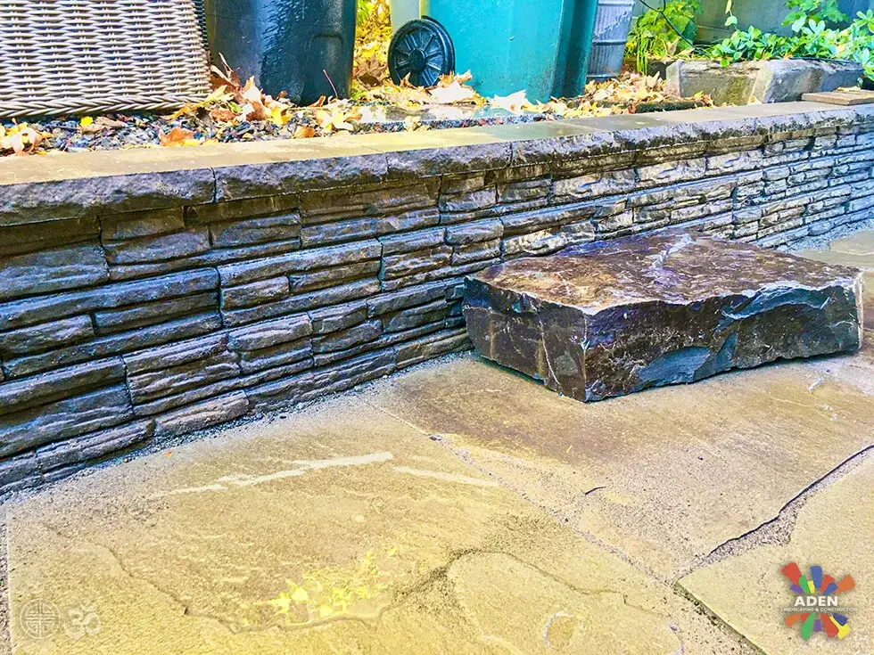 Wall of thin stacked grey stone with a single thick stone slab set in front of it as a bench, on a concrete terrace