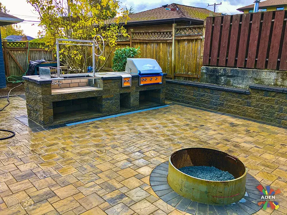 Outdoor kitchen with two built-in stainless grills on a stone-faced counter beside a paver patio, with a copper fire bowl set into the surface